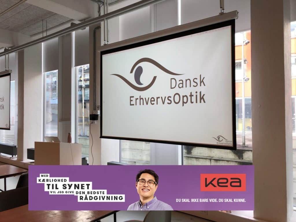 Projektorlærred med Dansk ErhvervsOptik-logo i lyst lokale, nederst banner med KEA-reklame og rådgiver.